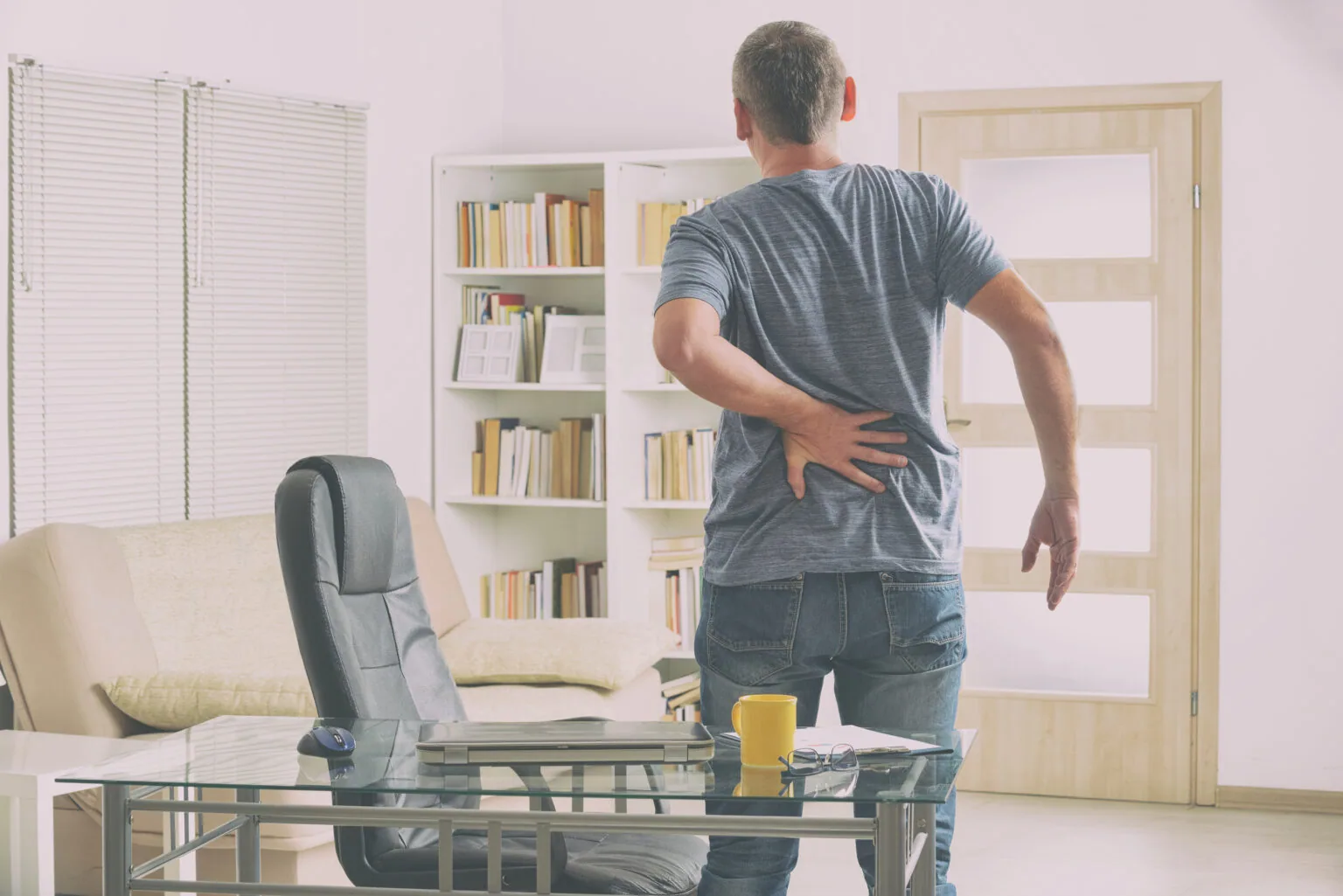 Man standing in office with hand on lower back, showing signs of back pain