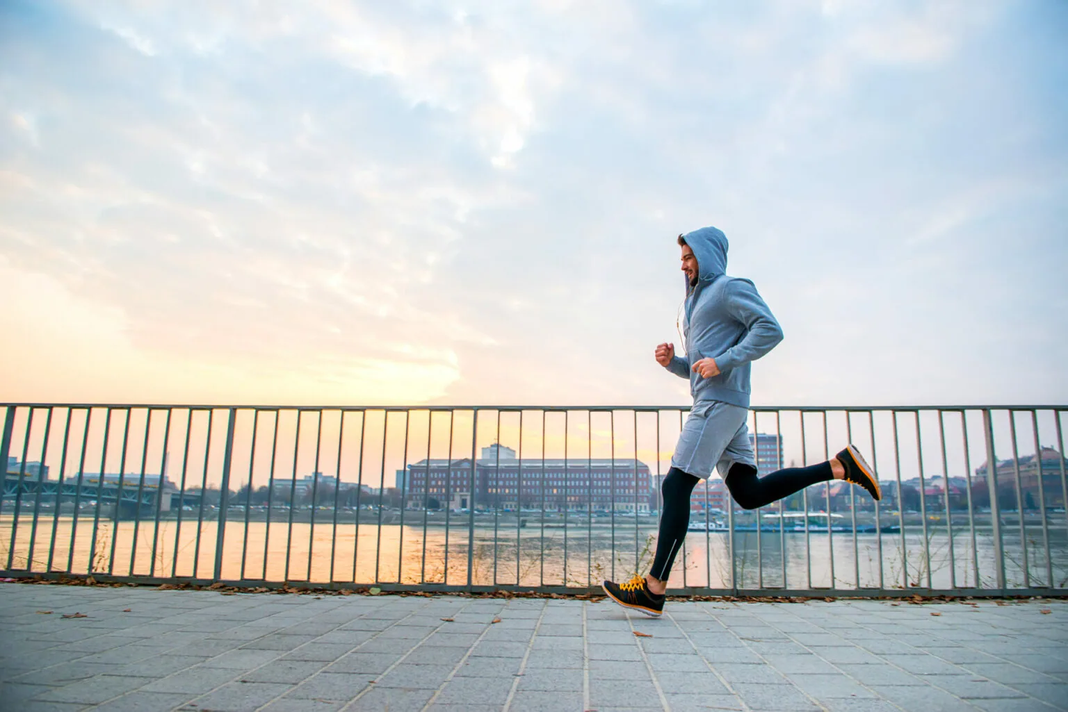 Person jogging in blue hoodie on paved waterfront path with metal railing and city skyline in background