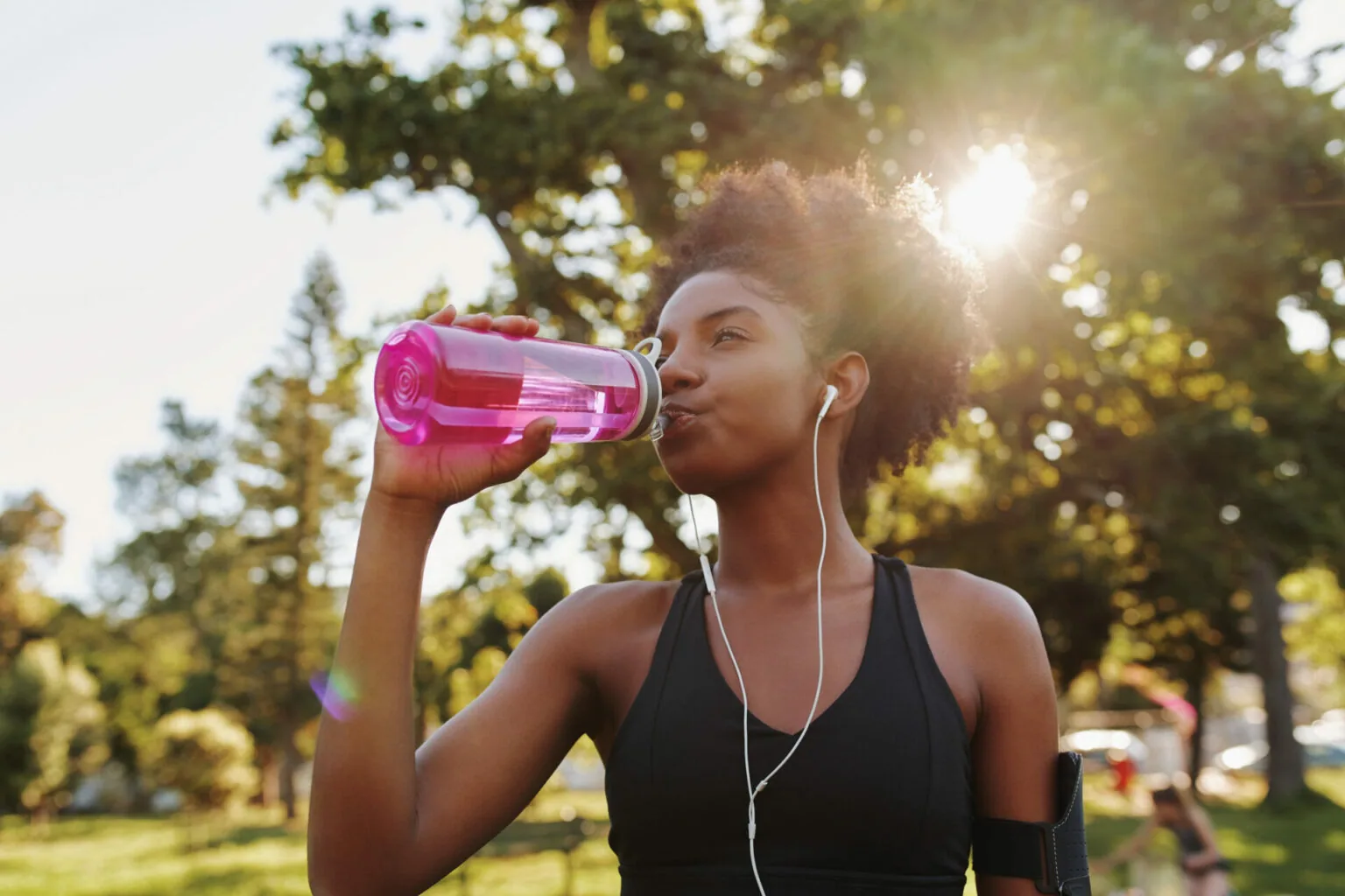 Woman in athletic wear drinking from pink water bottle outdoors in park with earbuds in