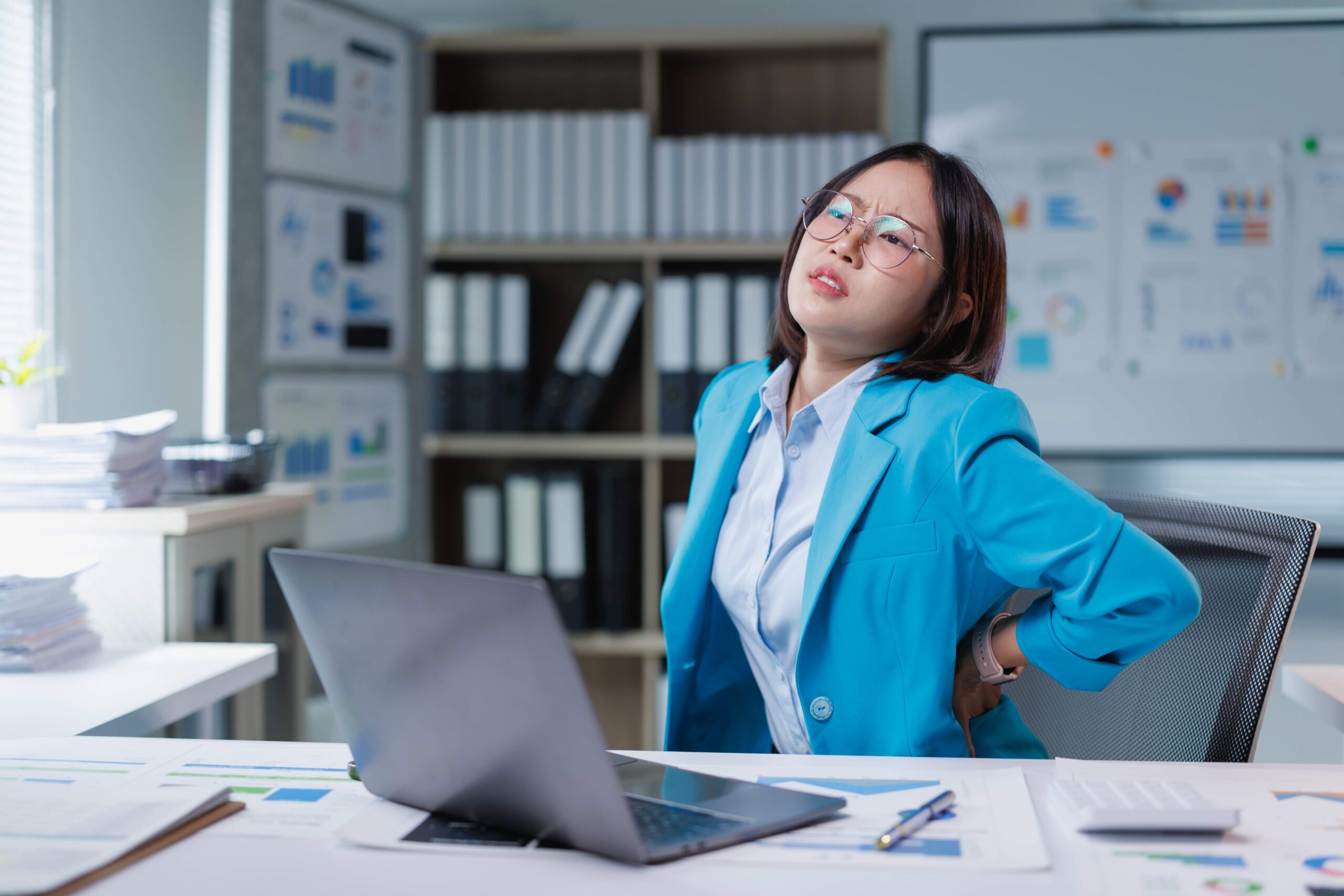 Woman in blue blazer sitting at office desk with laptop, holding lower back in discomfort while looking upward