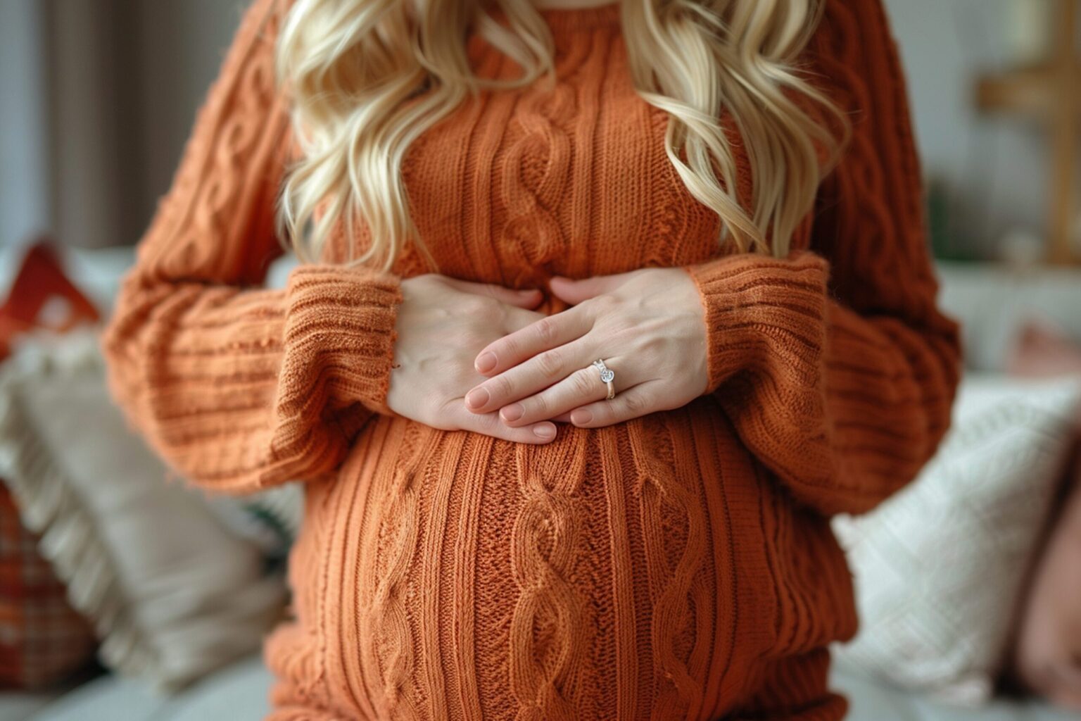 Pregnant woman in a rust cable-knit sweater cradling her baby bump with both hands, wearing a diamond engagement ring.