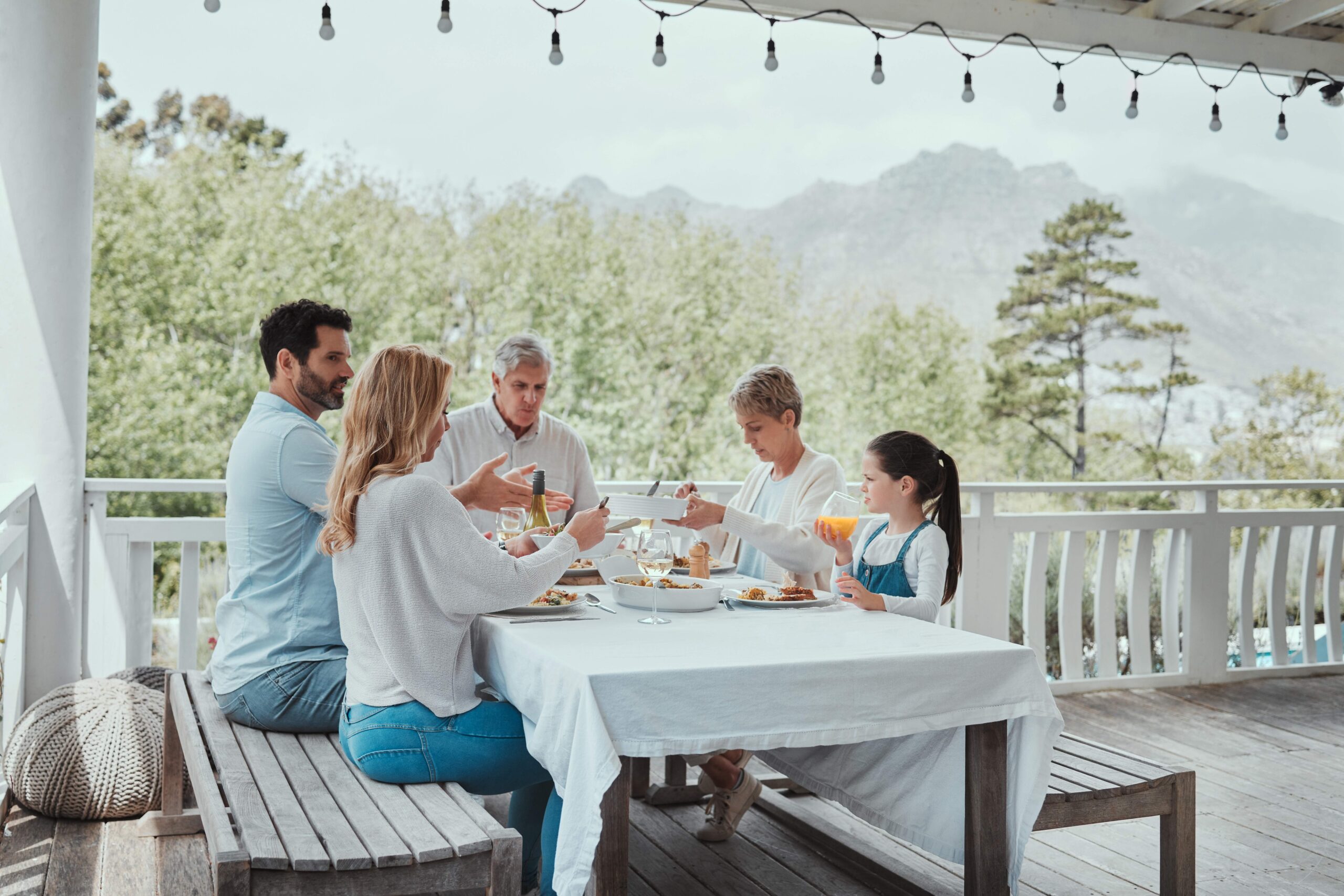 Five-person multigenerational family dining together on a sunny deck with mountain forest views, sharing a meal at a white-clothed table.