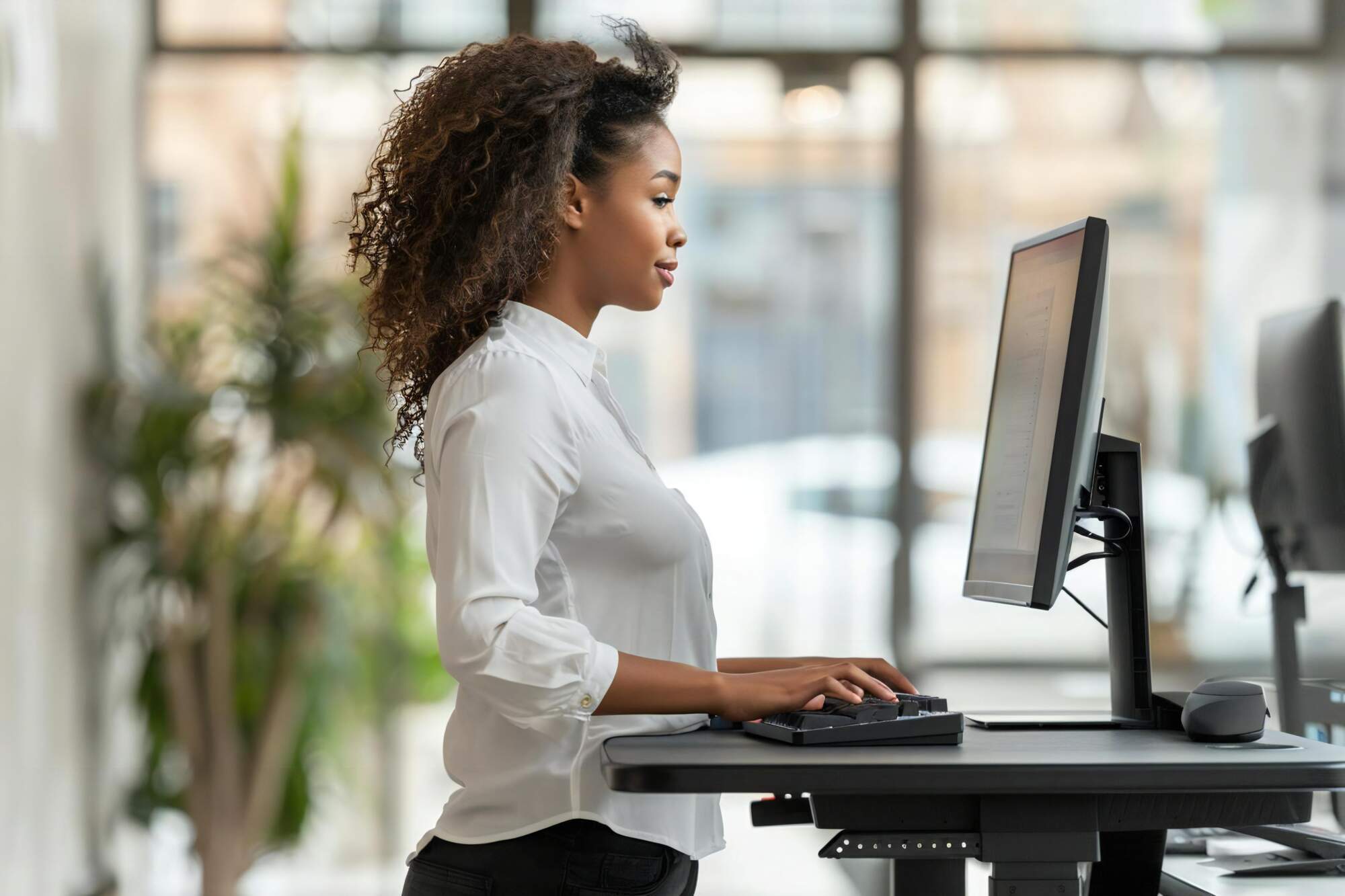 Woman sitting at desk with proper posture while working at computer in bright office with windows and plants