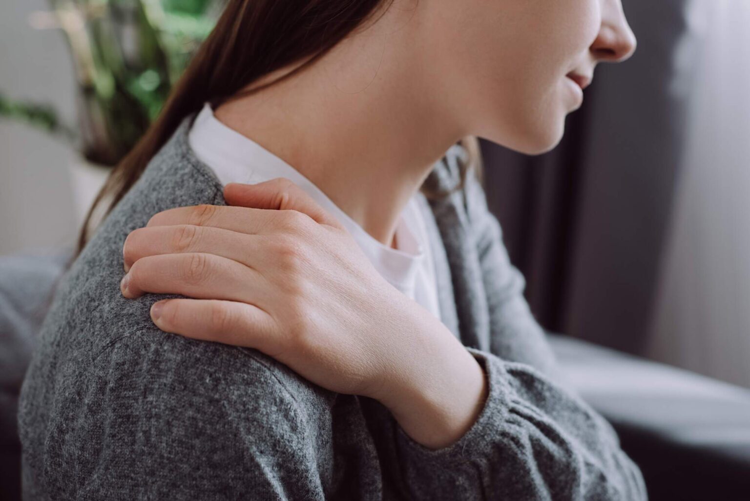 Woman in gray cardigan holding her shoulder, appearing to experience discomfort or stiffness