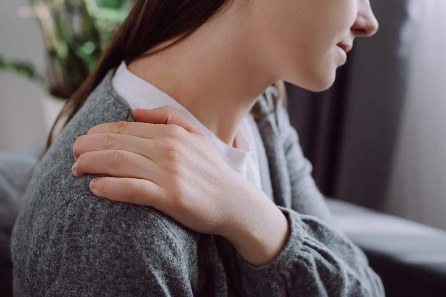 Woman in gray cardigan holding her shoulder, appearing to experience discomfort or stiffness