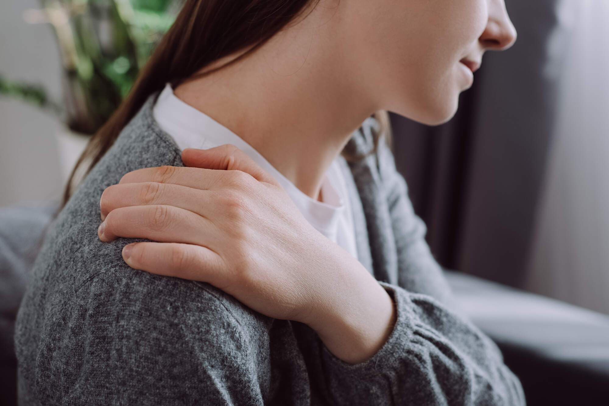 Woman in gray cardigan holding her shoulder, appearing to experience discomfort or stiffness