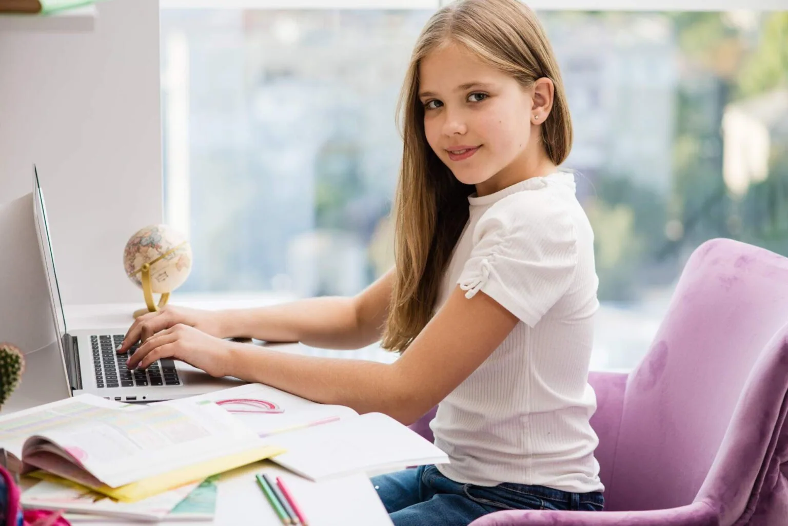 Young girl with blonde hair sitting at a desk with a laptop, globe, and study materials, looking over her shoulder at the camera with a slight smile