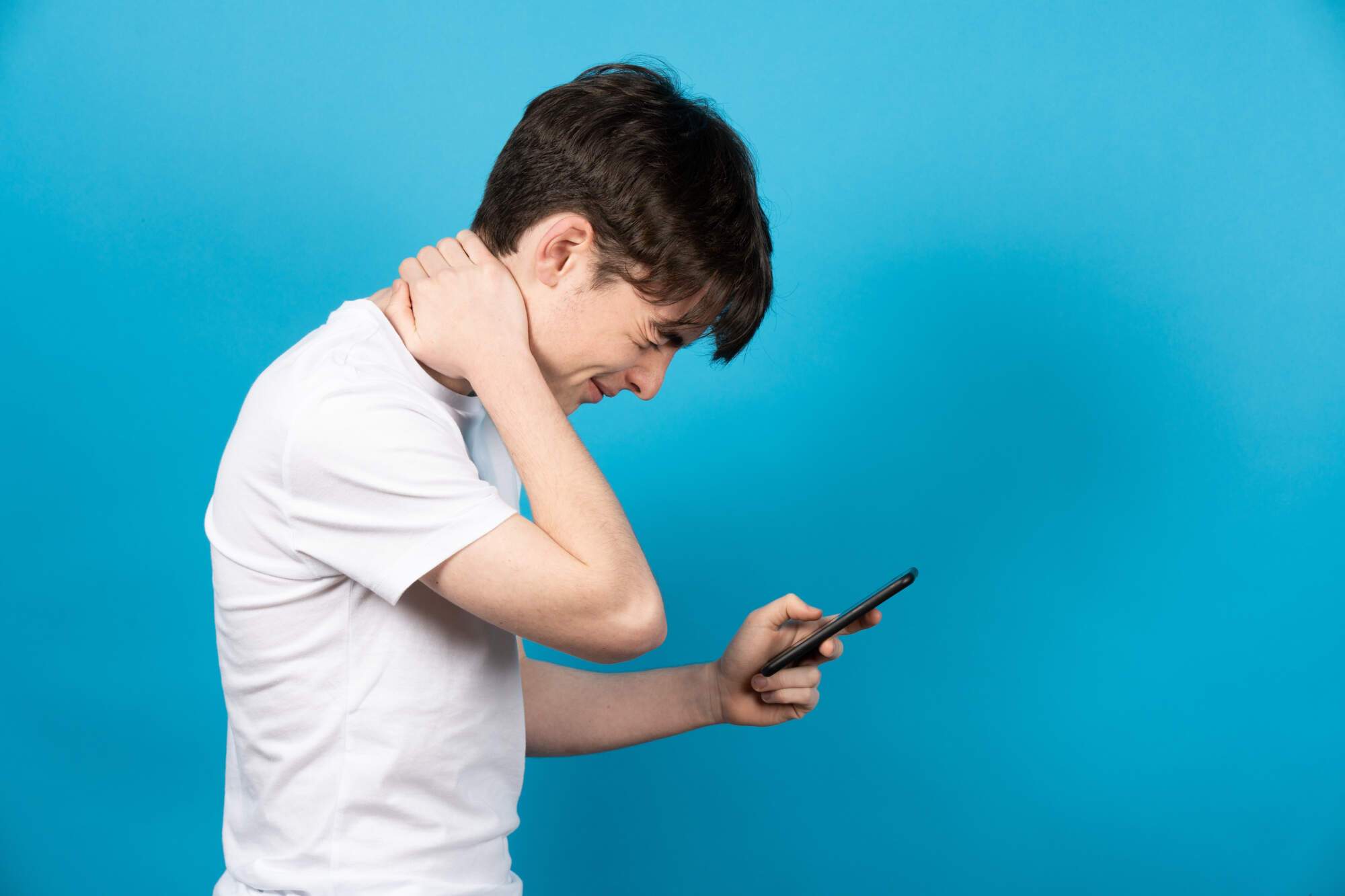 Young teen in white shirt holding smartphone, looking down with hand on neck showing discomfort against blue background